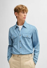 Ljusblå denimskjorta med långa ärmar, spetsig krage, två bröstfickor och knappframslutning. Bärs med beige byxor.