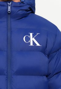 Blå pufjakke med høj krave og lynlås, der har et hvidt "cK" logo på venstre bryst.