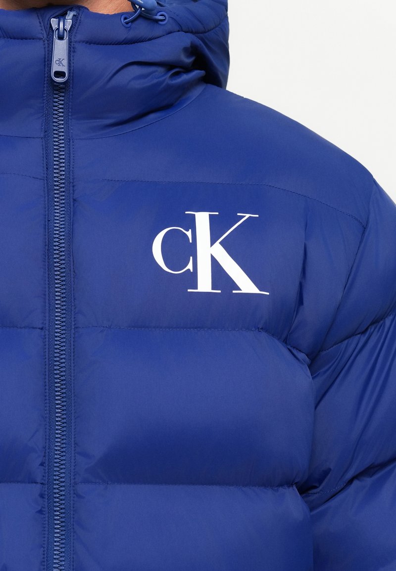 Kék puffertaki magas gallérral és cipzárral, fehér "cK" logóval a bal mellrészen.