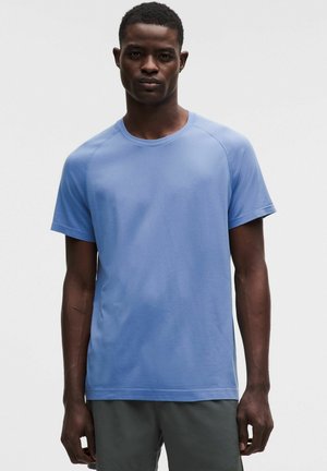 Man draagt een lichtblauw sportshirt met korte mouwen en donkergrijze shorts, staat tegen een effen lichte achtergrond.