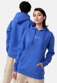 Modra bombažna kapuca s sprednjim žepom, kapuco na vezalko in izvezenim belim logotipom. Ima rebraste manšete in rob, mehka tekstura.