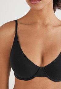 Černá podprsenka s tenkými ramínky, hladkým povrchem a hlubokým výstřihem. Nabízí tvarované košíčky a bezešvé okraje pro čistý vzhled.