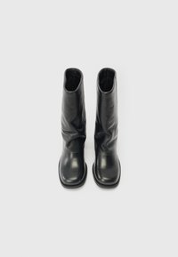 Par de botas de cuero negro de media caña con punteras redondeadas y un eje ligeramente arrugado, exhibidas en posición vertical sobre fondo blanco.