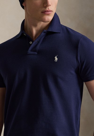 Joven con una camiseta polo azul marino con un pequeño logo blanco de jugador de polo en el pecho izquierdo, mirando ligeramente hacia la derecha.