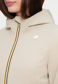 Beige capuchonjack met een kleurrijke rits met gele, blauwe en rode strepen. Inclusief een klein logo aan de voorkant. Glad textuur.
