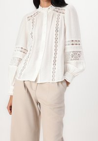 Valkoinen napillinen paita, jossa on pitsireunus, pitkät puffihihat ja röyhelökaulus. Paita on pukeutunut vaaleanbeigeihin housuihin, joissa on teksturoitu kangas.