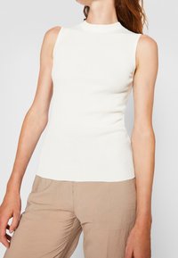 Mujer vistiendo una blusa sin mangas de canalé blanca y pantalones beige, contra un fondo claro y liso, con los brazos relajados a los lados.