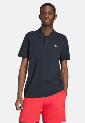 Młody mężczyzna w czarnej koszulce polo Adidas i czerwonych szortach Adidas, stoi z jedną ręką w kieszeni na jednolitym tle.