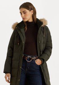Cappotto imbottito verde oliva con cappuccio foderato in pelliccia sintetica, dotato di tasche con cerniera e coulisse in vita. Indossato sopra un dolcevita scuro.