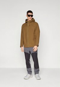 Καφέ αδιάβροχο μπουφάν με πράσινο φερμουάρ, γκρι παντελόνι με μαύρες λεπτομέρειες και γκρι αθλητικά παπούτσια. Το μοντέλο φοράει γυαλιά ηλίου.