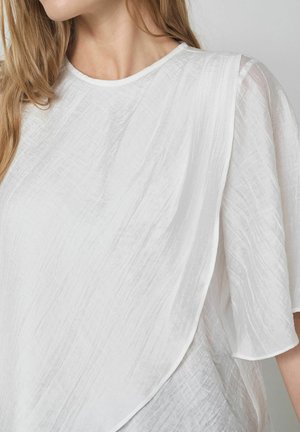 Vrouw draagt een witte blouse met textuur, korte wijd uitlopende mouwen en een ronde halslijn, bovenlichaam en nek zichtbaar.