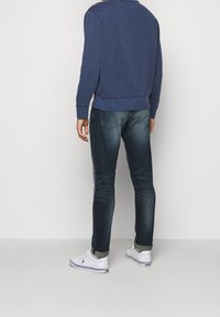 Man med blekta blå jeans, upplagda ärmar, vita sneakers och en långärmad marinblå sweatshirt, står mot en enkel vit bakgrund.