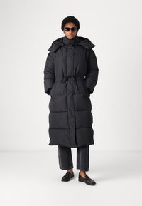 Črna puffer jakna z visokim ovratnikom, z žABC-om v pasu, dolgimi rokavi in dolžino do kolen. Obuta je v temne jeans hlače in črne čevlje.