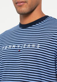 Sinine ja tumet sinine triibuline T-särk ümmarguse kaelusega ning valge "TOMMYJEANS" logo. Logo all on väike punane, valge ja sinine aktsent.