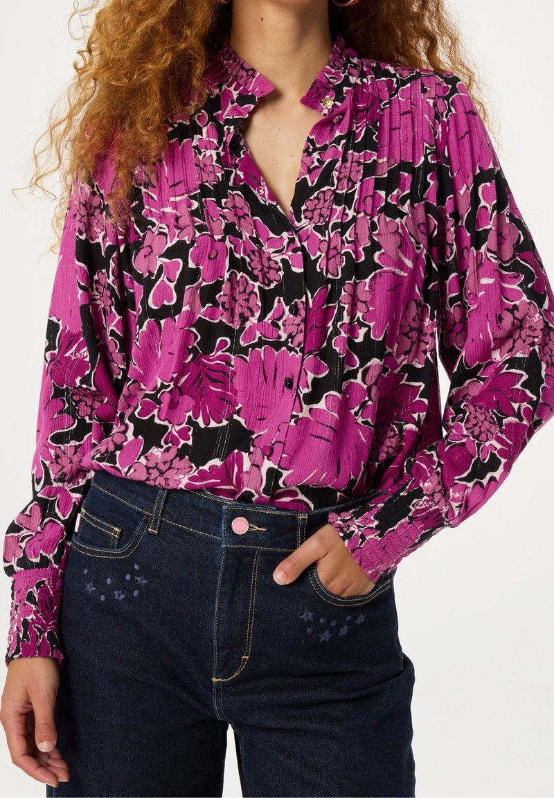 Femme portant un chemisier à manches longues à motif floral rose, noir et blanc, rentré dans un jean bleu foncé, main dans la poche.