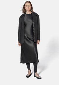 Robe en satin noir, silhouette ajustée, longueur genoux, associée à un manteau noir. Les chaussures présentent des détails décoratifs sur des ballerines pointues.