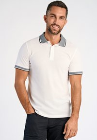 Vit polo shirt med grå kontrasterande krage och ärmslut, utrustad med två knappar och en slim fit-design, kombinerad med mörka byxor.