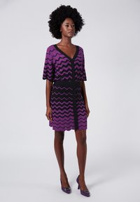 Abito a maniche corte in viola e nero, con motivo a zigzag, scollo a V e bottoni decorativi sulla parte frontale. Texture liscia.