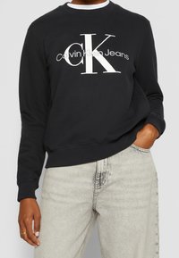 Black sweatshirt with white "Calvin Klein Jeans" logo. Cotton blend, crew neckline, ribbed cuffs and hem, worn with light grey jeans.