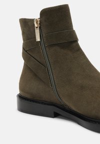 Bottine en suède vert olive avec semelle noire, fermeture éclair latérale avec tirette dorée, et détail de sangle autour de la cheville.