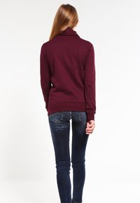 Jersey burdeos con cuello alto, tejido suave, diseño ajustado y puños de canalé, combinado con jeans azul oscuro con bolsillos traseros.