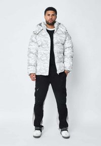 Doudoune camouflage blanche, dotée d'une capuche, d'une fermeture éclair et d'une texture matelassée. Associée à un pantalon cargo noir et des baskets blanches.