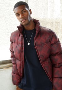 Rød mønstrede pufferjakke med en blank finish, med høj krave og lynlås, båret over en navy blå t-shirt og sølv halskæde.