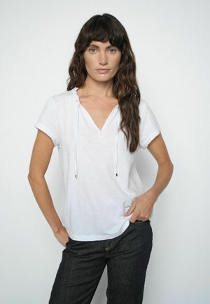 Woman with long wavy dark hair wearing a white short-sleeve V-neck top with ties and black pants, standing with one hand in pocket.