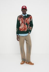 Πλεκτό πουλόβερ σε σκούρο πράσινο με μεγάλο πολύχρωμο σχέδιο, φορεμένο πάνω από ένα ανοιχτό μπλε πουκάμισο με γιακά, συνδυασμένο με μπεζ παντελόνι φαρδιάς γραμμής και καφέ αθλητικά παπούτσια.