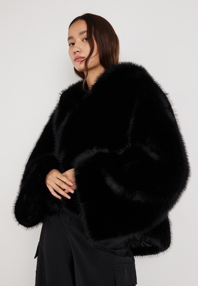 Juoda dirbtinio kailio striukė su tūrinėmis rankovėmis, minkšta tekstūra ir plačiu apykakliu. Turi lygią apdailą ir laisvą siluetą.