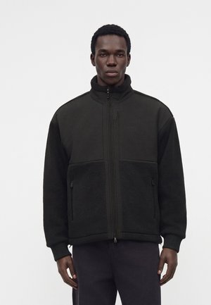 Homme portant une veste polaire noire zippée avec un col montant et des poches zippées, debout devant un fond clair uni.