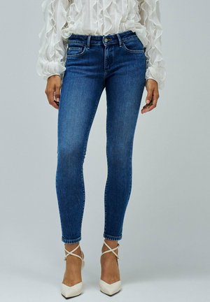 Jean skinny en denim bleu avec une finition délavée, taille mi-haute et détails subtils sur les poches avant, associés à des talons blancs à lanières.