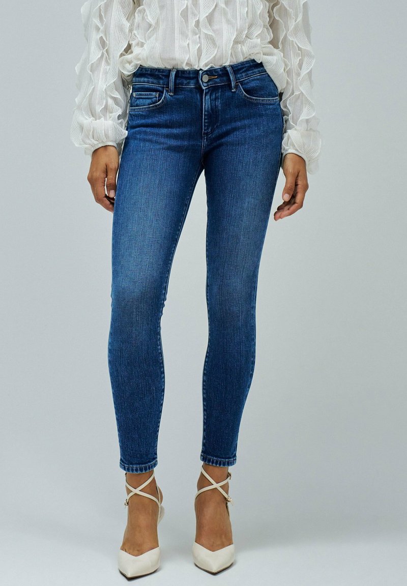 Jeans de denim azul skinny con un acabado desgastado, cintura de altura media y detalles sutiles en los bolsillos frontales, combinados con tacones blancos con tiras.