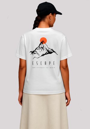 Frau, die ein weißes T-Shirt mit einem Berg- und roten Sonnen-Graphic sowie dem Wort "ESCAPE" auf dem Rücken trägt, kombiniert mit einem beigen Rock und einer schwarzen Kappe.