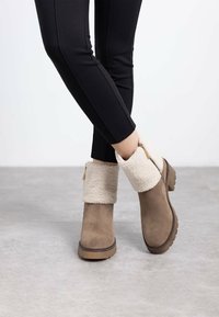 Botas de tobillo de ante marrón con un puño de piel sintética color crema y detalle de cremallera dorada, con una suela texturizada y un diseño elegante.