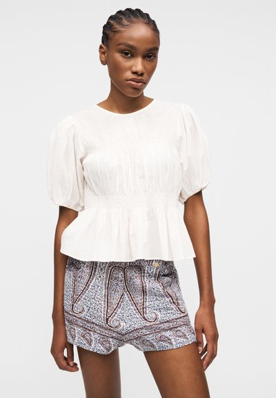 Femme aux cheveux tressés portant un chemisier blanc à manches bouffantes et un short à motif paisley, debout devant un fond clair uni.