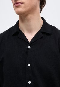 Νεαρό άτομο που φοράει μαύρο πουκάμισο με κουμπιά και γιακά, εμφανίζοντας το πάνω μέρος του στήθους και του λαιμού σε απλό φόντο.