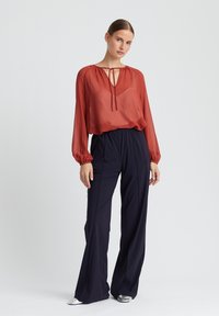 Transparente rostfarbene Bluse mit Schlüsselloch- und Schleifendetail am Ausschnitt, kombiniert mit hochgeschnittenen, marineblauen, weit geschnittenen Hosen und silbernem Schuhwerk.