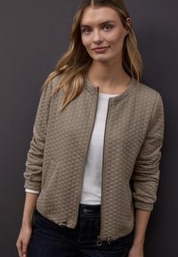 Gesteppte beige Bomberjacke mit rundem Halsausschnitt, durchgehendem Reißverschluss vorne und seitlichen Taschen. Wird über einem weißen Top getragen und hat gerippte Bündchen und Saum.