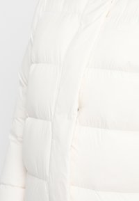 Chaqueta tipo puff en blanco roto, con secciones acolchadas, cierre frontal de cremallera y una textura suave. Diseño ligero para mayor calidez.