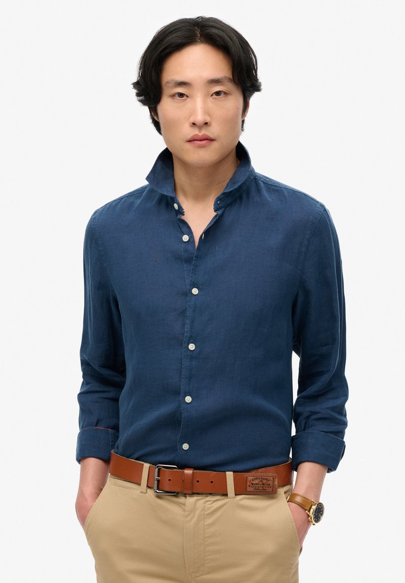 Marineblauwe linnen overhemd met een hoge kraag, knoopsluiting, en opgerolde mouwen, gedragen met beige broeken en een bruine riem.