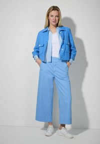 Lichtblauw cropped jasje met twee voorkruiszakken, knoopdetails en lange mouwen over een witte top. Gecombineerd met wijd uitlopende lichtblauwe broek en witte sneakers.
