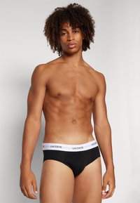 Schwarze Baumwollslips mit einem weißen elastischen Bund, der mit dem Lacoste-Logo versehen ist. Modelliert von einem Mann mit lockigem Haar und einem durchtrainierten Körper.