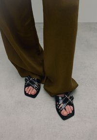 Zwarte sandalen met straps en vierkante tenen, voorzien van metalen oogjes en een glanzende afwerking, gecombineerd met olijfgroene wijde broek.