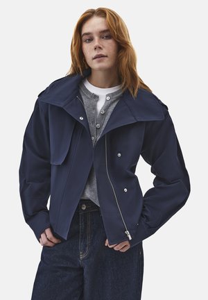 Młoda kobieta z rudymi włosami, ubrana w granatową kurtkę na zamek, szary kardigan na guziki, białą koszulę i ciemnoniebieskie dżinsy, stojąca z rękami blisko kieszeni.