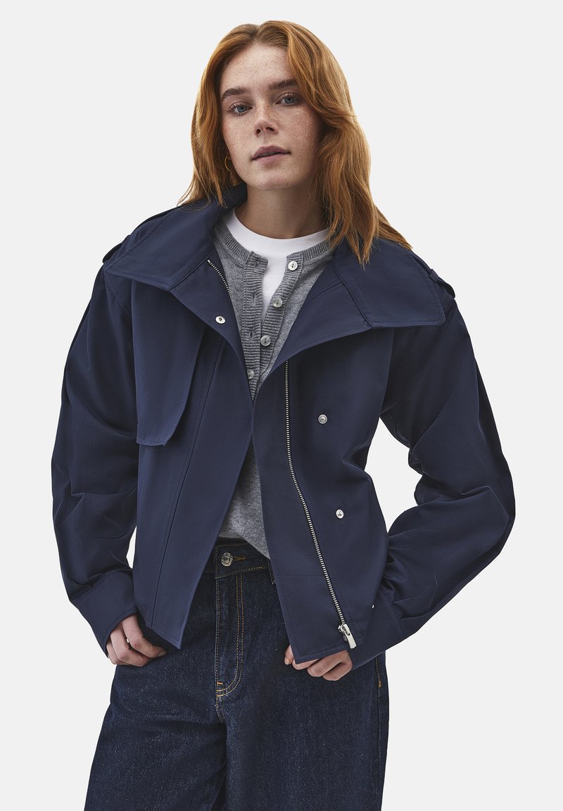 Jonge vrouw met rood haar, gekleed in een marineblauwe jas met ritssluiting, een grijs vest met knopen, een wit shirt en donkere blauwe jeans, staand met handen nabij haar zakken.