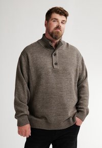 Uomo con barba che indossa un maglione lavorato a maglia marrone con bottoni e pantaloni neri, in piedi con una mano in tasca su uno sfondo semplice.