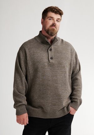 Mann mit Bart, der einen braunen, geknöpften Strickpullover und schwarze Hosen trägt, steht mit einer Hand in der Tasche vor einem einfarbigen Hintergrund.