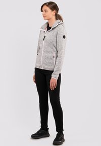 Grau melierte Fleecejacke mit Reißverschluss und Kapuze, kontrastierendem pinken Reißverschluss, schwarzen Akzenten und zwei Fronttaschen. Getragen mit schwarzen, engen Hosen.