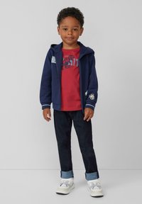 Junger Junge steht, trägt einen navyfarbenen Reißverschluss-Hoodie, ein rotes T-Shirt mit Dinosauriergrafik, dunkle hochgekrempelte Jeans und weiße Sneakers, neutraler Hintergrund.